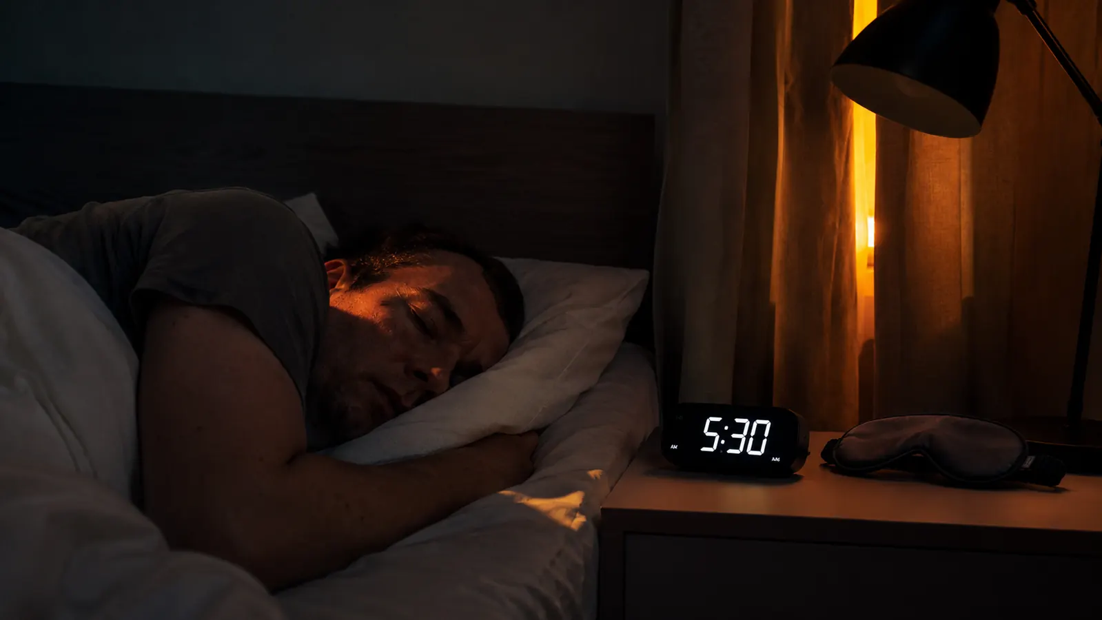Dark bedroom at 5:30am with street light visible through window gap and a sleep mask on the nightstand